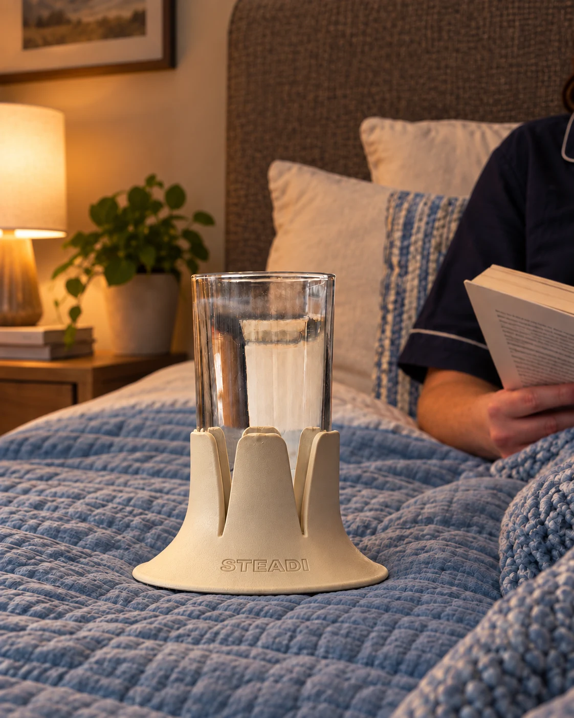 A water glass in a Sand-colored Steadi on blue bedding beside an adult reading. Reference-guided AI lifestyle image.